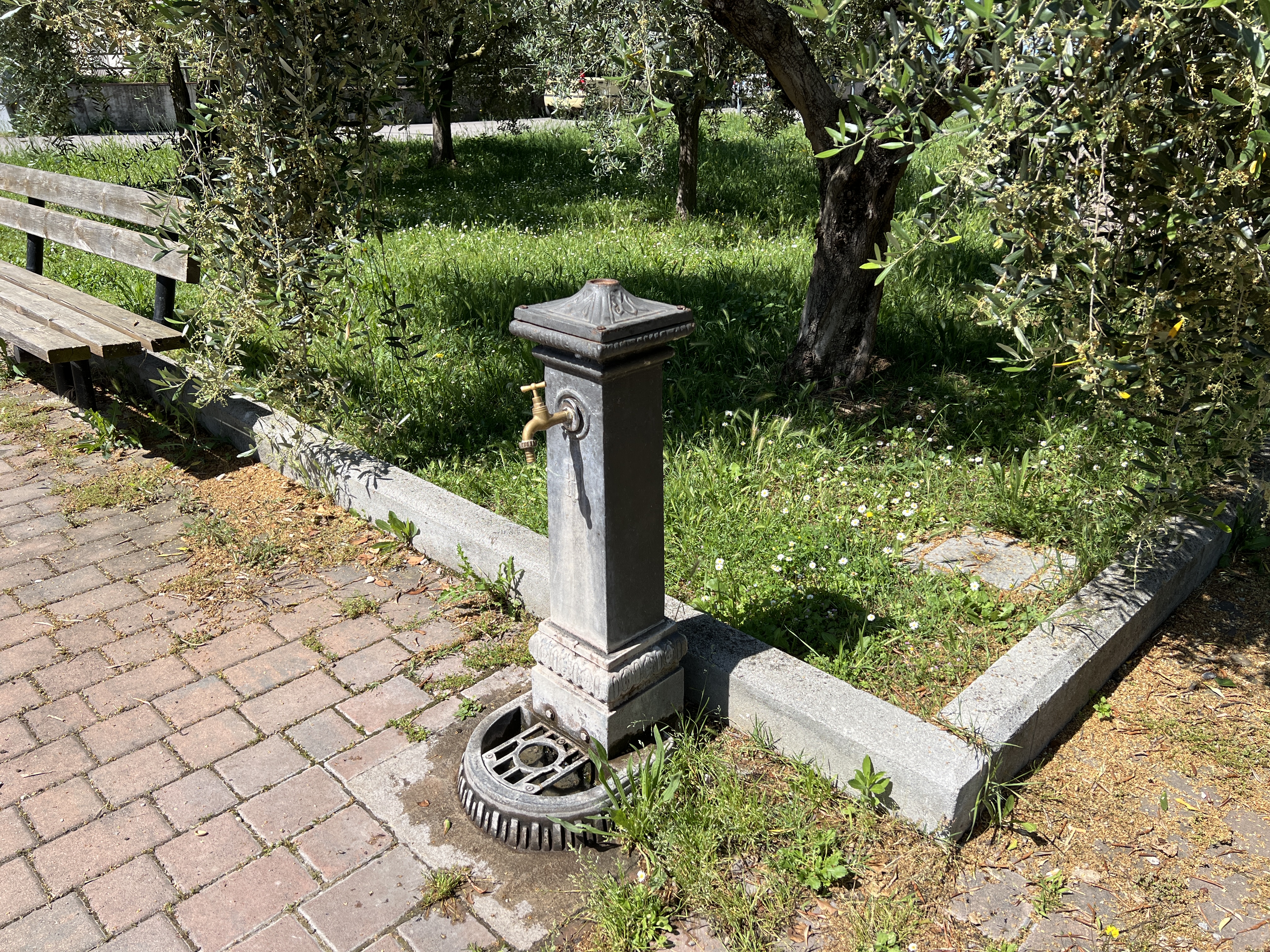 Metal drinking fountain with gold tap in Torricella park. Red brick pavement. Green area and bench.